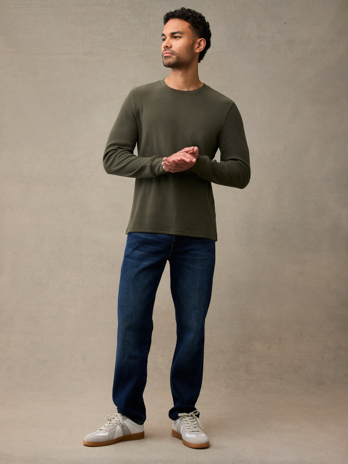 Man wearing an army green long-sleeve flannel shirt, dark blue jeans, and white sneakers, standing. Layers clean under flannel, fleece, or a shell jacket