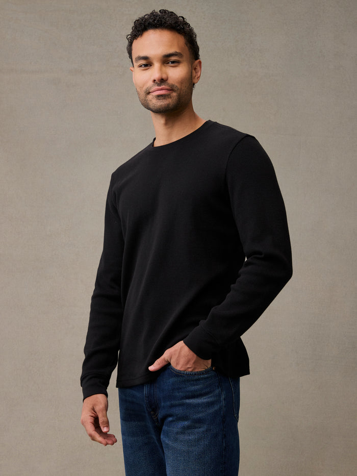 A man wearing a black long-sleeve flannel and blue jeans, standing against a textured gray studio background. Upgraded fabric: softer, thicker waffle, better shape retention.