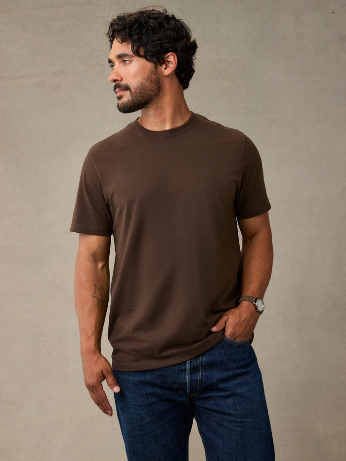 A man wearing a coffee brown short-sleeve crewneck t-shirt and blue jeans, standing against a textured gray wall. Heavyweight enough for the job, comfortable enough for after.