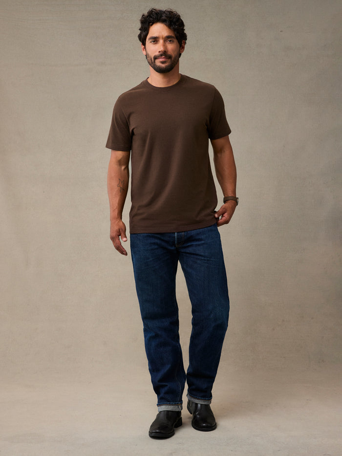A man wearing a brown short-sleeve crewneck t-shirt, dark blue jeans, and black shoes. The tee that holds up to the whole weekend's to-do list.