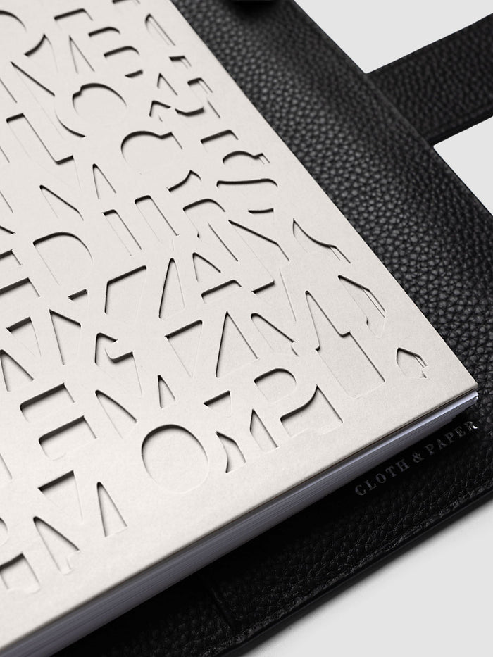 Close-up of the A5 6-Ring Punch Diecut Pocket Folder at an angle on a black pebbled leather cover, showing the depth of the cut letters and the printed logo along the edge of the solid back panel