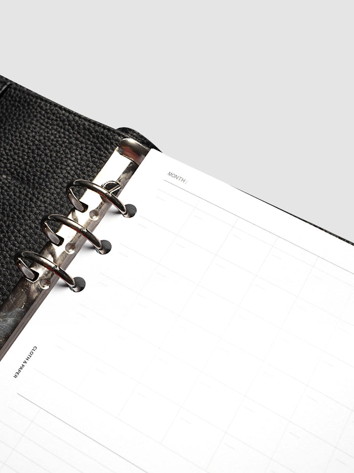Monthly Fold Out Planner Insert Set of 2, A5 6-Ring Punch, angled close-up of the top left corner showing the month line, the gray calendar grid, and the silver rings against black pebbled leather