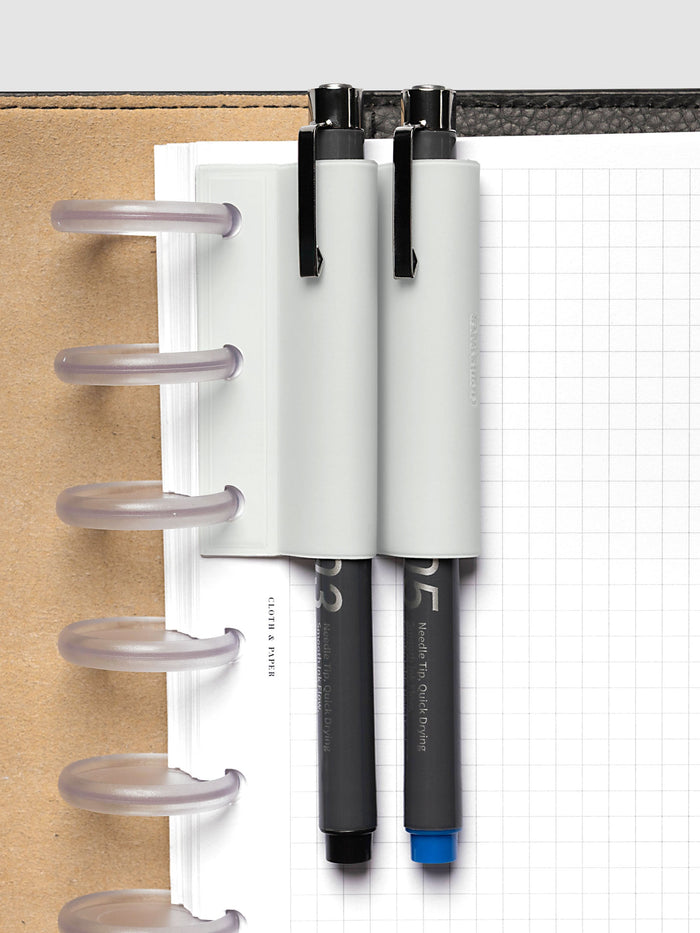 System Punch Pen Holder | Aspen, disc punch, pale gray holder carrying two gray barreled drawing pens on frosted clear discs above a white grid page