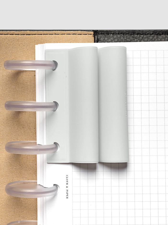 System Punch Pen Holder | Aspen, disc punch, pale gray holder seated on frosted clear discs in a black leather planner over a white grid page