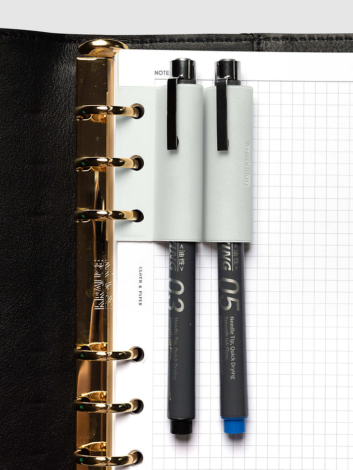 System Punch Pen Holder | Aspen, ring punch, pale gray holder on gold rings holding two gray drawing pens, one capped in black and one capped in blue, over a white grid page