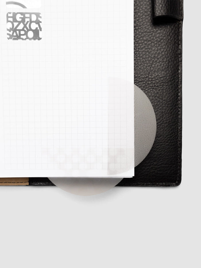 A Transparent Semicircle Sticky Note in use on the edge of a graph paper page in a black leather planner, extending past the page as a tab